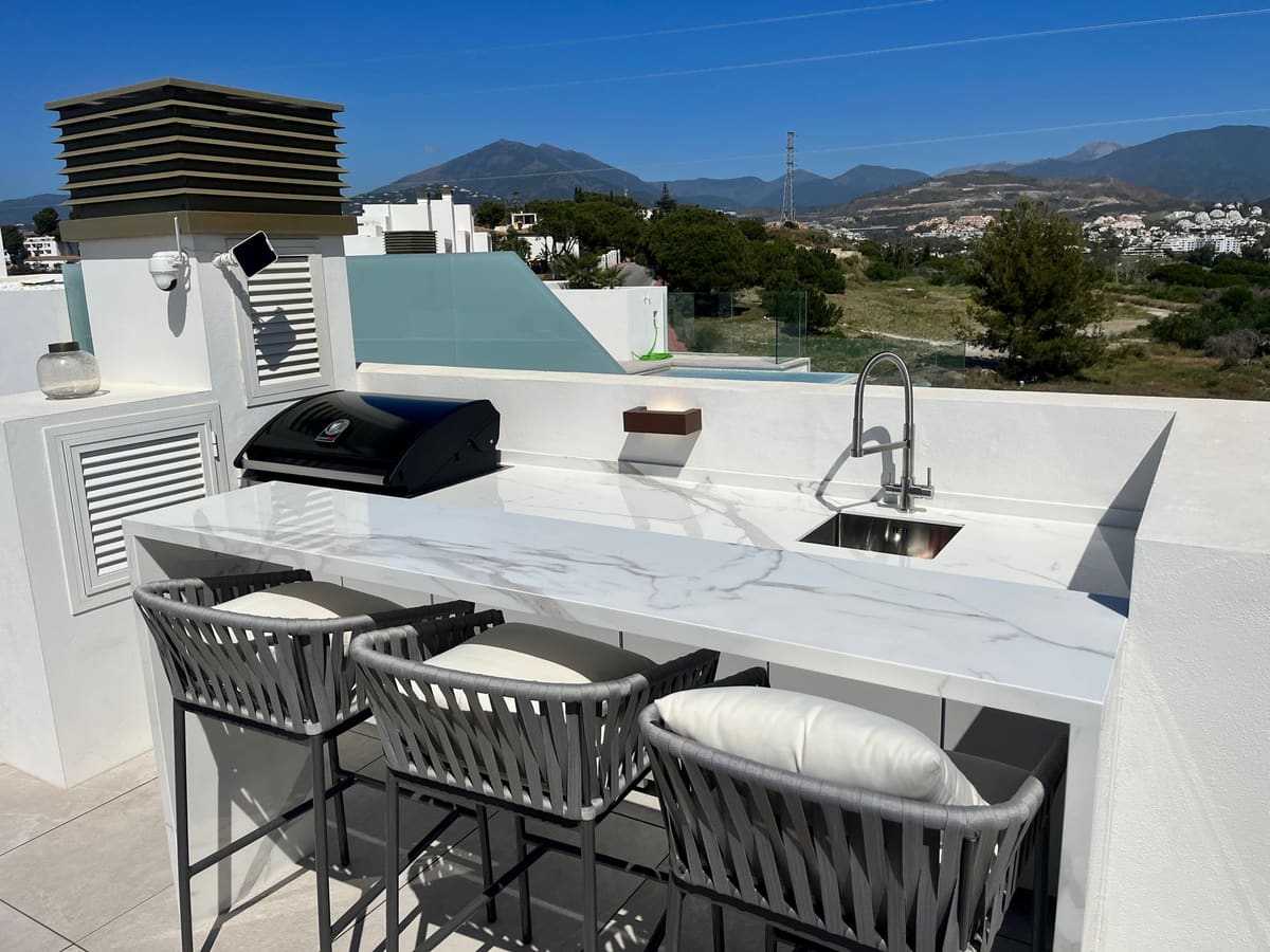 Rooftop outdoor kitchen with marble-effect worktop, sink, BBQ and three cushioned bar stools facing the mountains